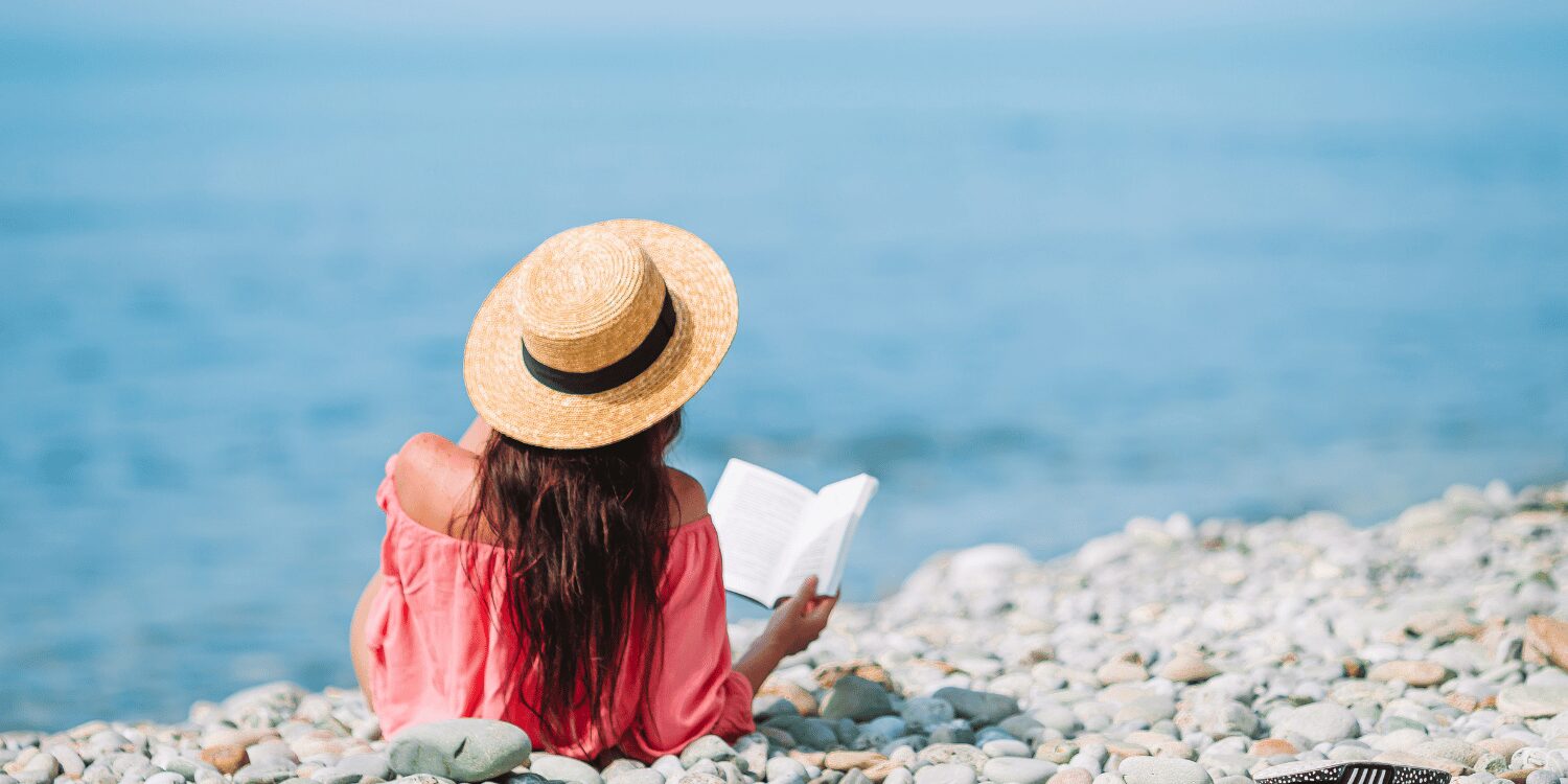 Frau mit Sonnenhut liegt auf Steinstrand und liest ein Buch.